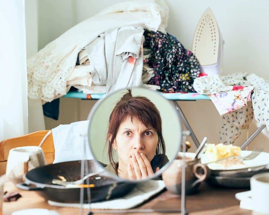 Woman looks with horror and fright at herself in mirror against background of mess